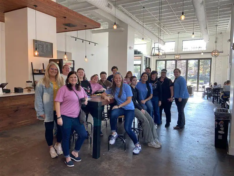 A group of smiling people in casual attire gather inside a bright, modern cafe with high ceilings and large windows.