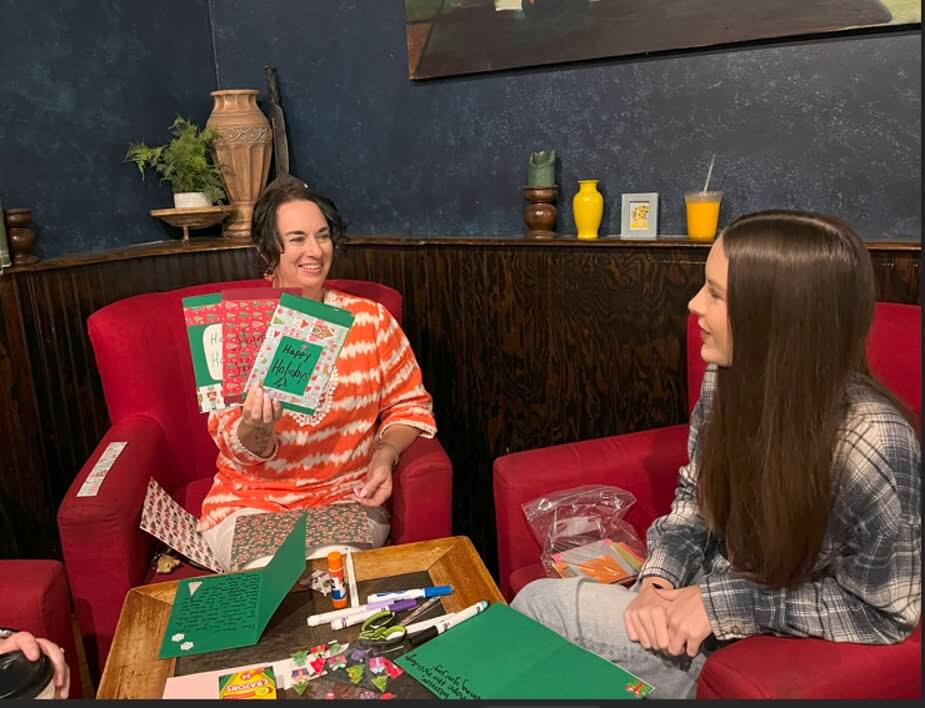 Two women sit in red chairs, smiling while crafting holiday cards.