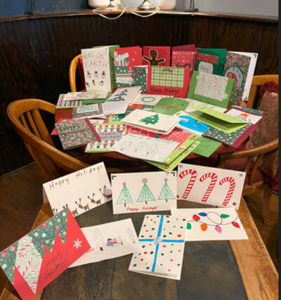 A wooden table is filled with colorful handmade holiday cards featuring festive designs like trees, candy canes, and lights.