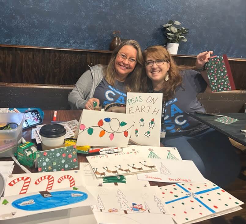 Two women smiling at a table covered with handmade holiday cards