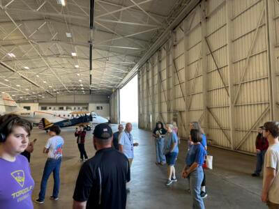 A group of people stand inside a spacious aircraft hangar with an airplane in the background.