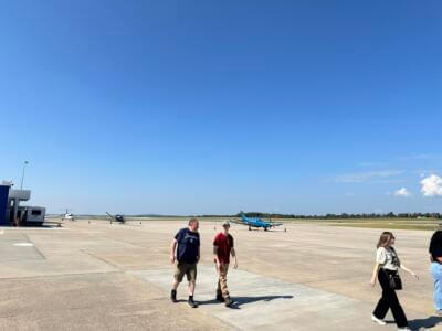 People walking on a sunny airport tarmac with small airplanes in the background.