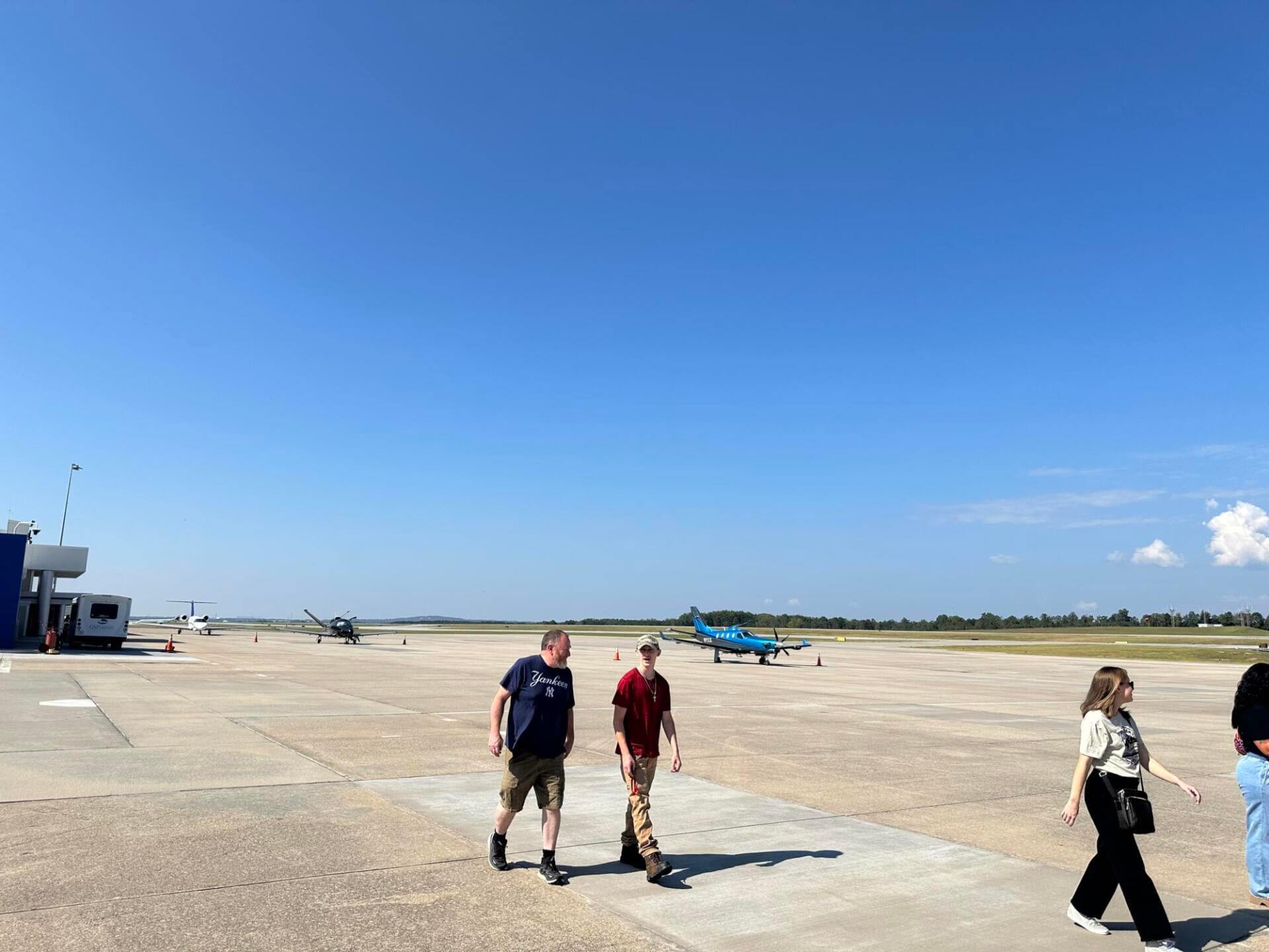 People walking on a sunny airport tarmac with small airplanes in the background.