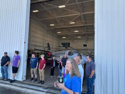 A group of people stand outside an open hangar with a small plane inside.