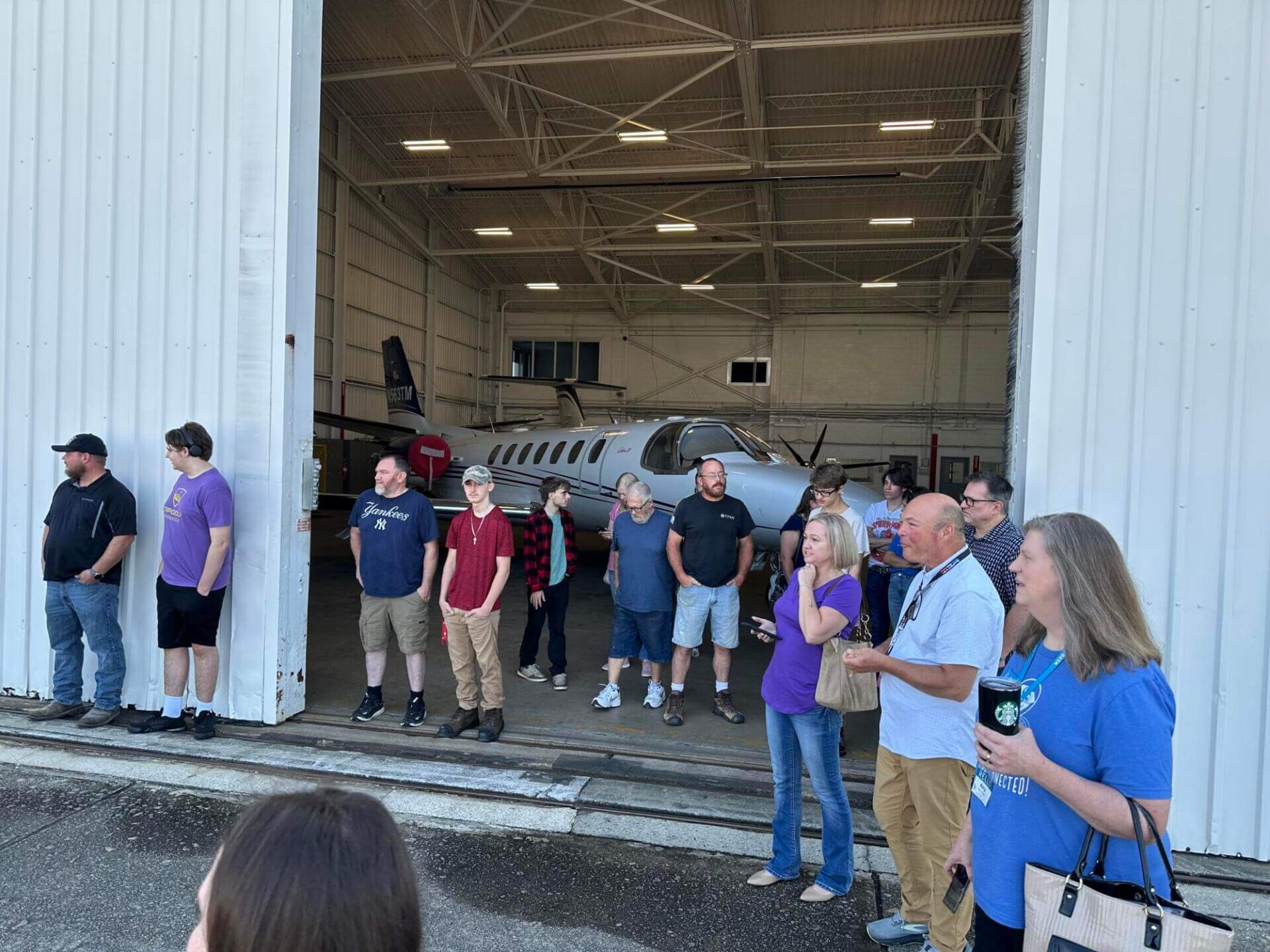 A group of people stands outside an open hangar door, inside which a small aircraft is partially visible.