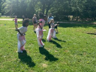 Four children are joyfully participating in a sack race on a sunny day in a grassy park. Onlookers watch as the kids hop in unison.