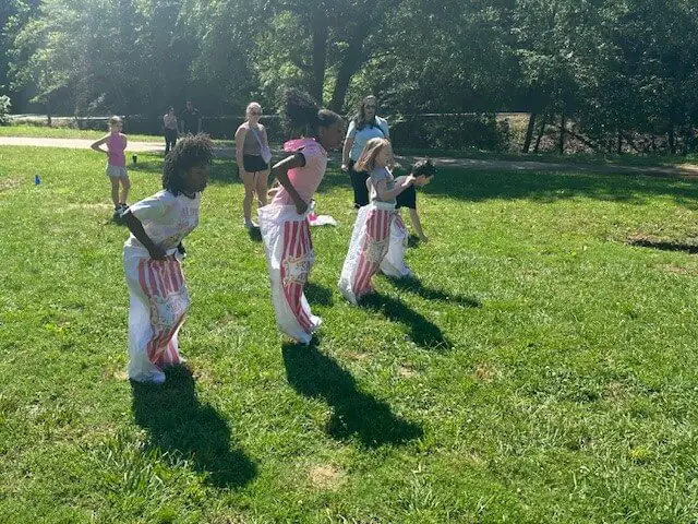 Four children are joyfully participating in a sack race on a sunny day in a grassy park. Onlookers watch as the kids hop in unison.