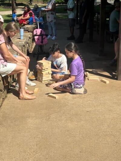 Two children play with large wooden blocks on a sunny path. They are focused and engaged. Nearby, adults and children relax on a stone bench.