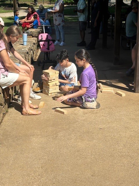 Two children play with large wooden blocks on a sunny path. They are focused and engaged. Nearby, adults and children relax on a stone bench.