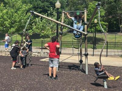 Children are playing on a modern playground structure in a park. One child sits on a swing, while others climb and hang on the metal framework.