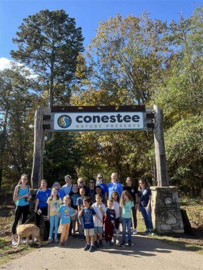 A group of people, including children and adults, stand happily beneath a sign reading "Conestee Nature Preserve,