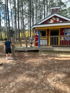 Two kids explore a colorful, small schoolhouse with a porch, set in a sunny forest with tall pine trees.