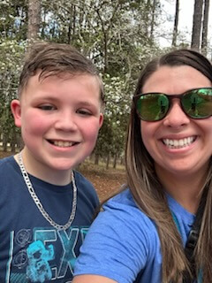 A smiling woman in sunglasses and a boy with a chain necklace pose outdoors