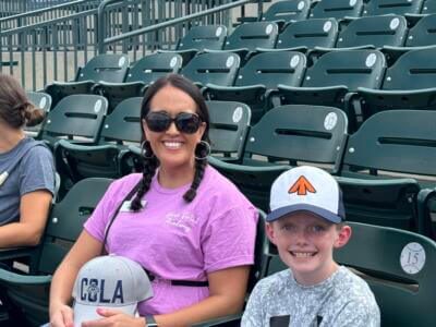 A woman in a purple shirt and sunglasses with braided hair smiles while sitting next to a child in a white cap.