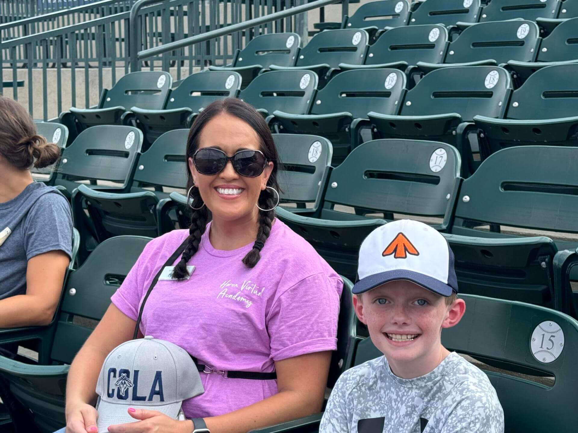 A woman in a purple shirt and sunglasses with braided hair smiles while sitting next to a child in a white cap.