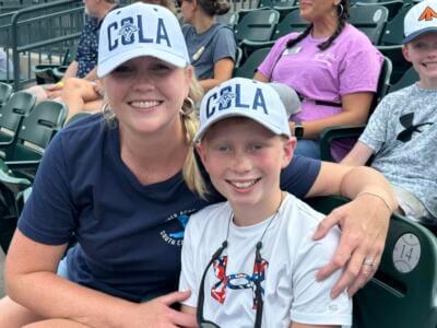 A smiling woman and child wearing matching "COLA" caps sit closely on stadium seats.