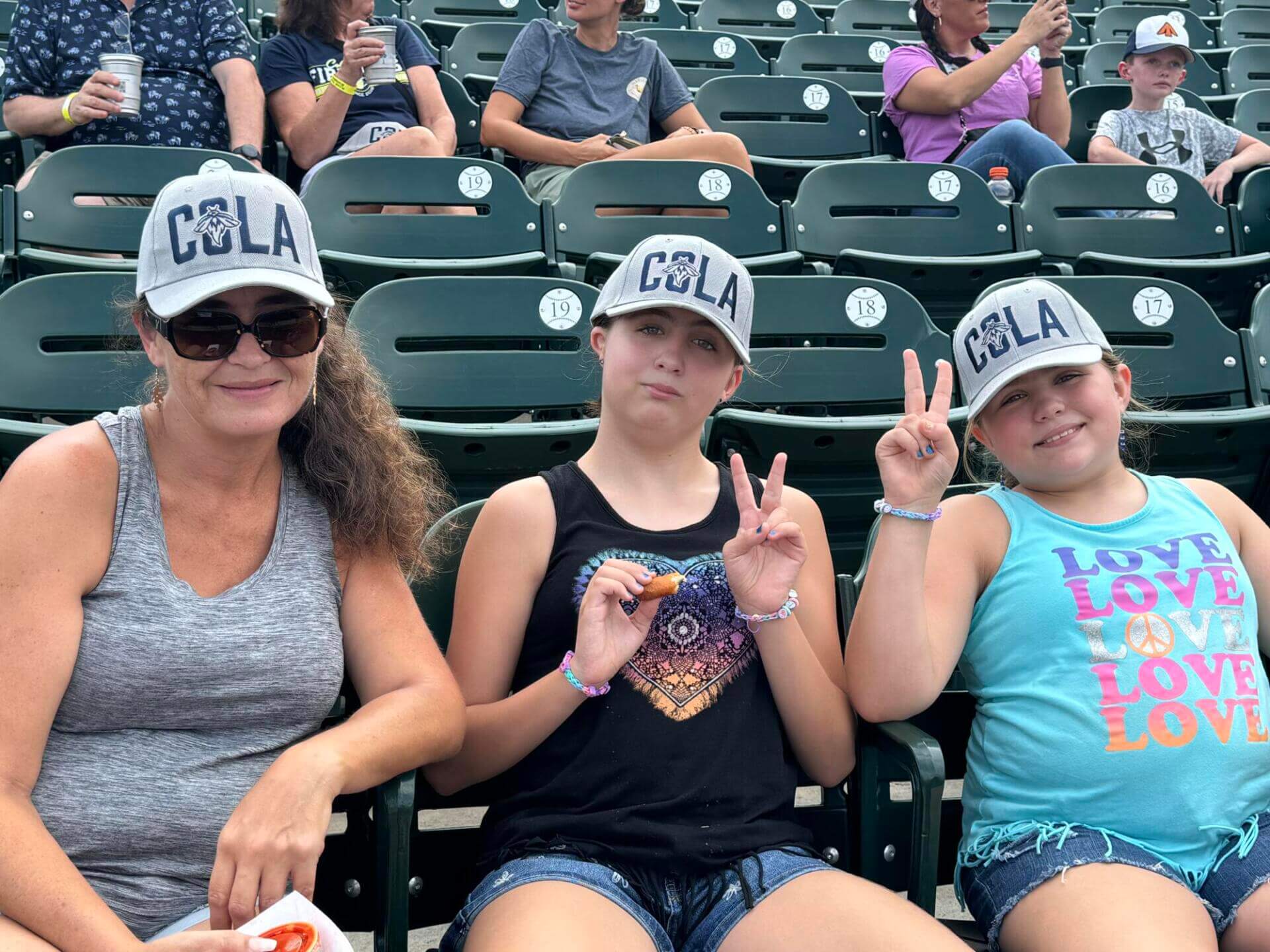Three people wearing "COLA" caps sit in a stadium.