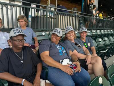 A group of four people sits in stadium seats, smiling and wearing matching "COLA" caps and blue shirts.