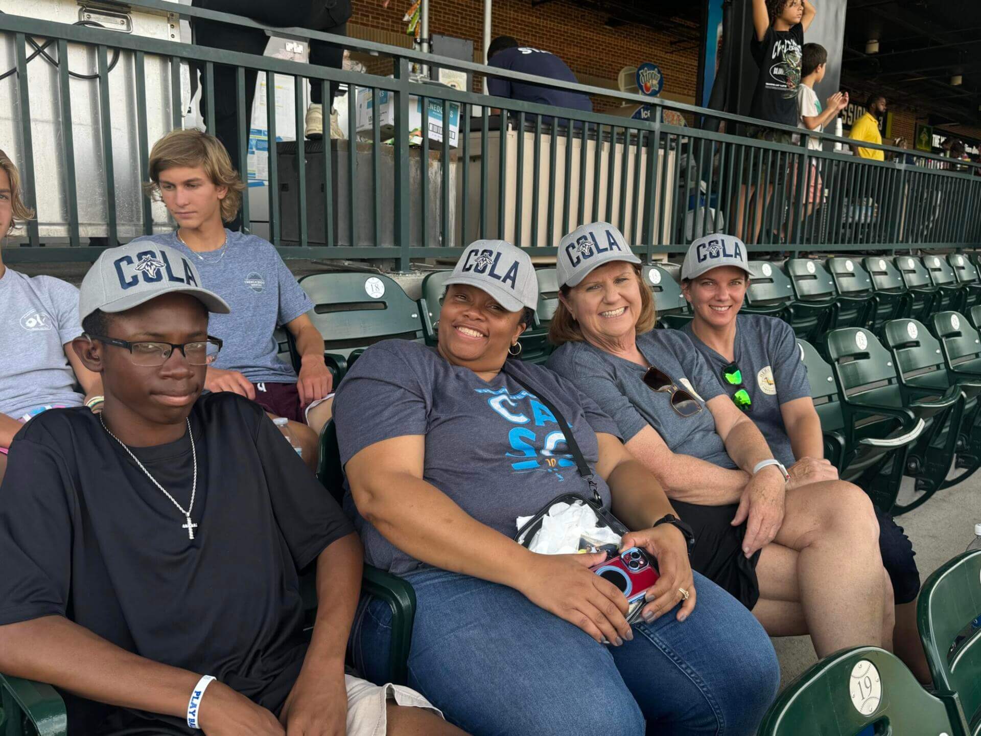A group of four people sits in stadium seats, smiling and wearing matching "COLA" caps and blue shirts.