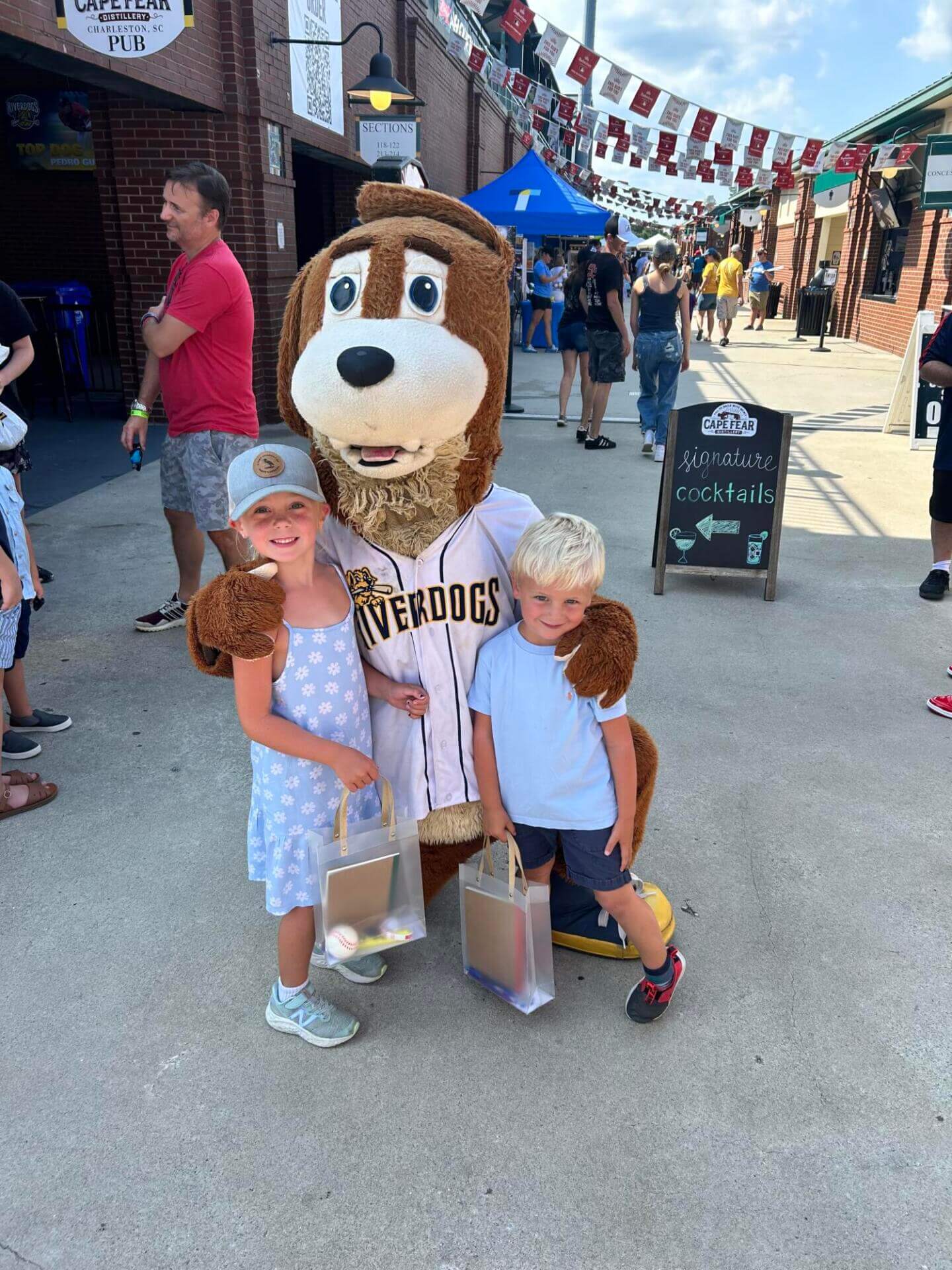 Two smiling children pose with a friendly dog mascot in a baseball jersey.