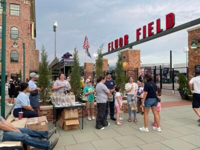 A diverse group of people gathers near the entrance of Fluor Field, enjoying a casual outdoor event.