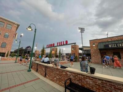 Outdoor scene at Fluor Field, featuring people gathering by brick structures and lampposts under a cloudy sky.