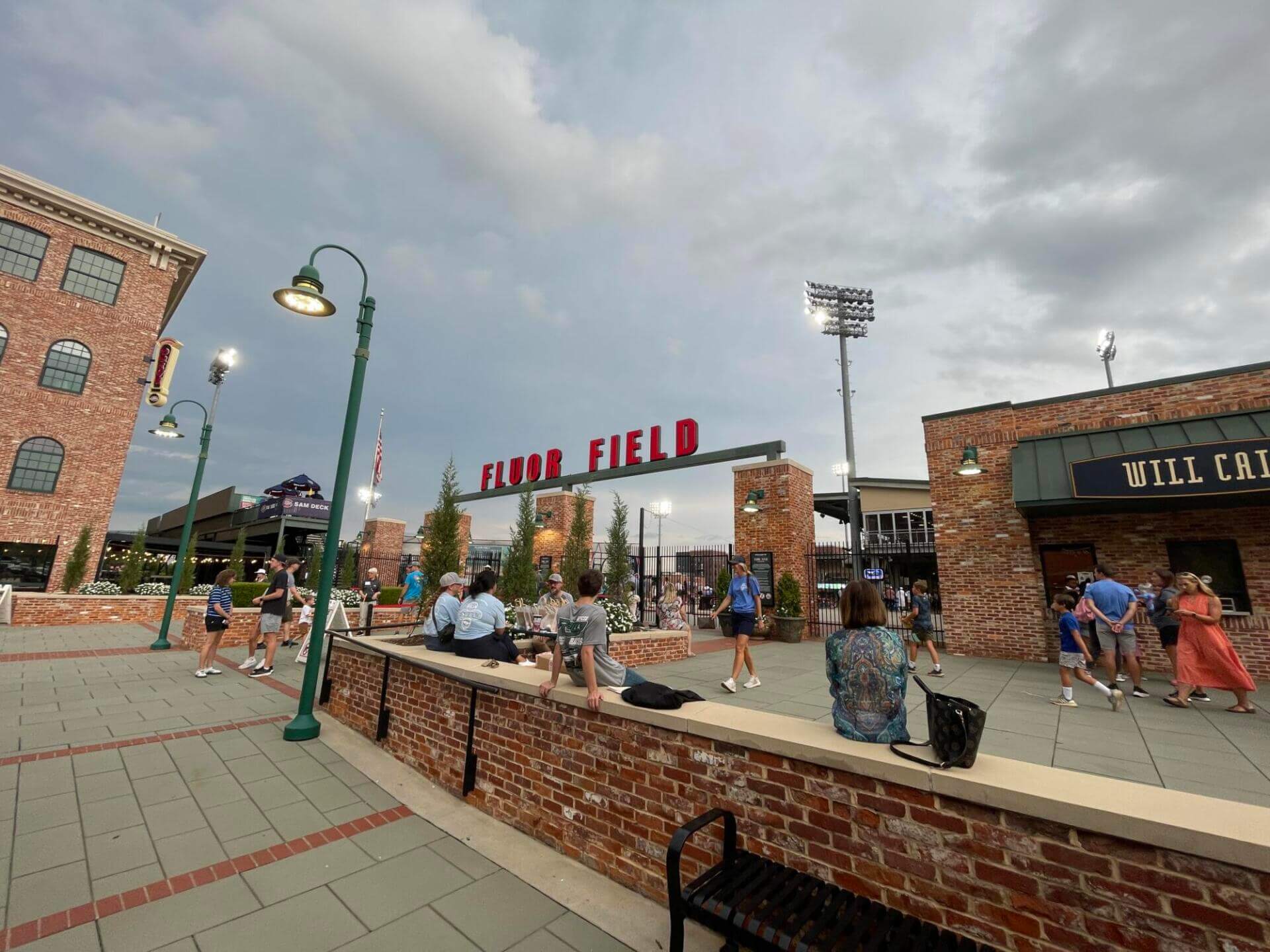 Outdoor scene at Fluor Field, featuring people gathering by brick structures and lampposts under a cloudy sky.