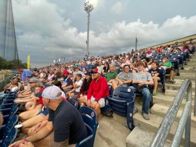 A large crowd sits in stadium seats under a cloudy sky, watching a sporting event.