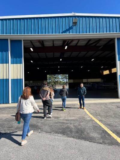 A group of people walk toward a large blue and white hangar under a clear blue sky.