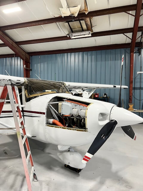 A small white aircraft inside a hangar, with an exposed engine and a propeller.