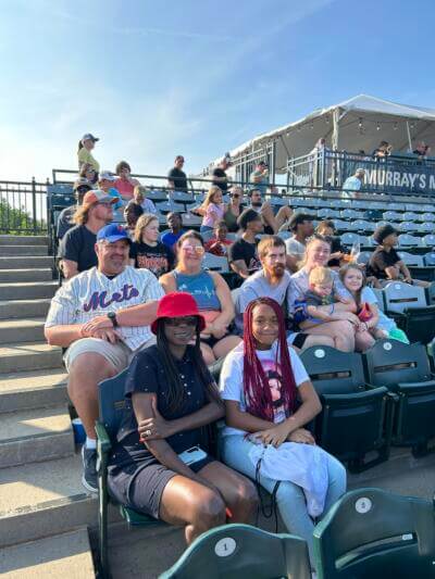 A group of people, including adults and children, sit in stadium seats on a sunny day.