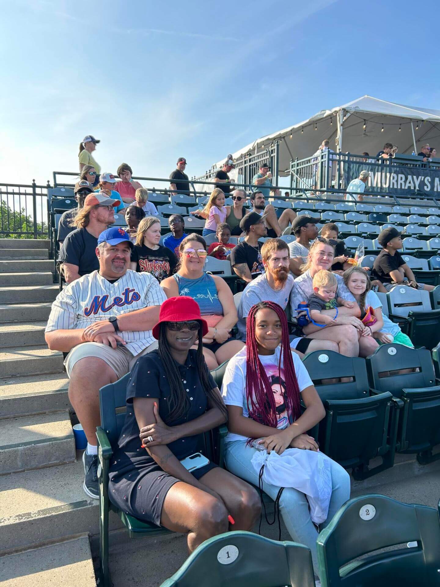 A group of people, including adults and children, sit in stadium seats on a sunny day.