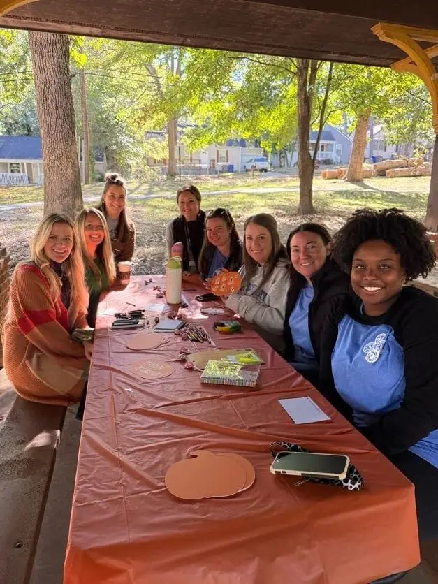 Group of smiling adults sits at an outdoor table covered with an orange tablecloth, crafting paper pumpkins.