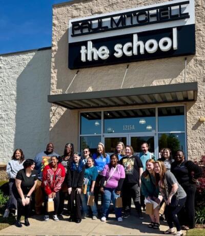 A diverse group of 18 smiling people stands in front of the "Paul Mitchell the School" building.