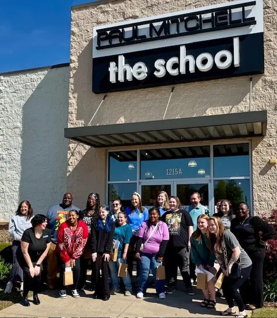 A diverse group of 18 smiling people stands in front of the "Paul Mitchell the School" building.
