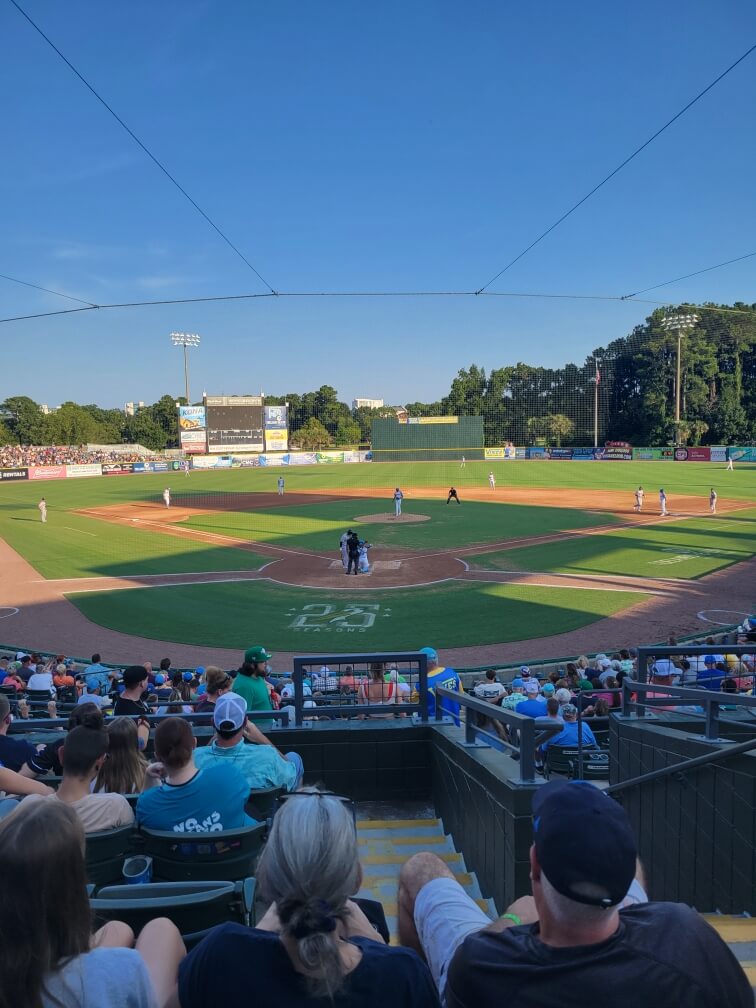 Baseball game in progress on a sunny day. Fans seated in a stadium watch players on the field.