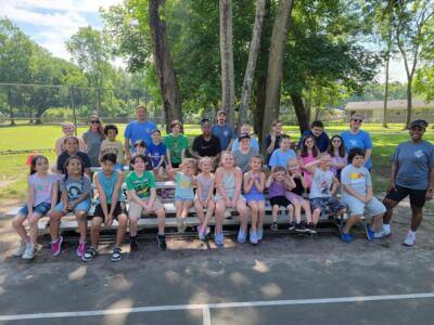 A diverse group of smiling children and adults sit on bleachers in a sunny park