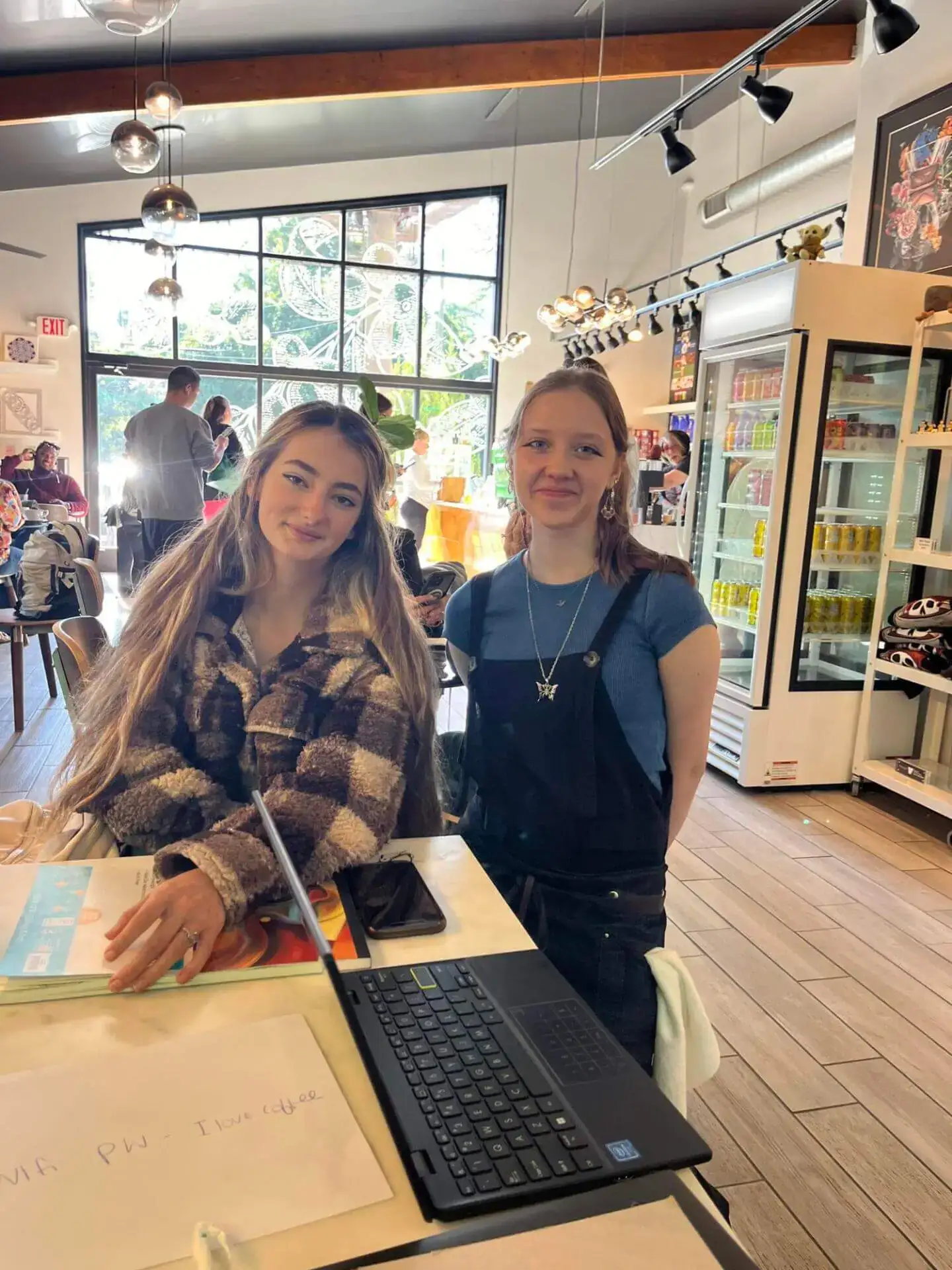Two young women smiling inside a cozy cafe with wooden decor and pendant lights.