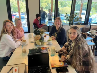 Four people smile around a café table with coffee and a laptop open