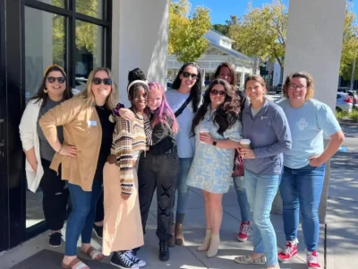 A group of eight smiling people stands outdoors on a sunny day near a building.