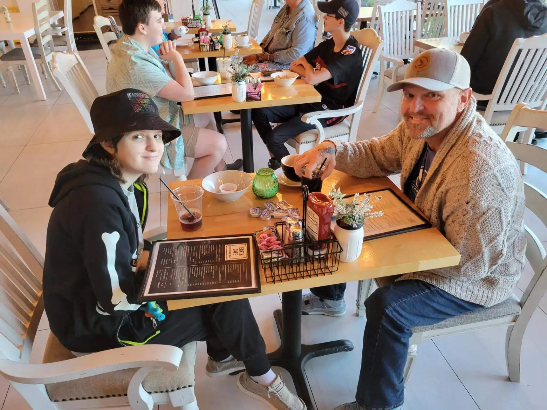 A person wearing a bucket hat and another in a cap smile at a café table with menus.