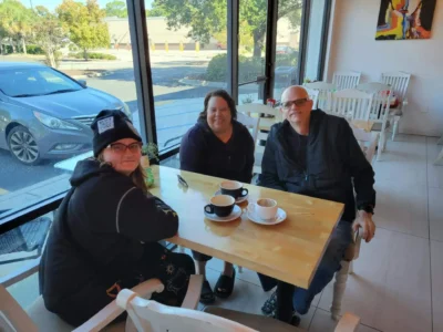 Three people sit at a wooden table in a cozy cafe by a large window.