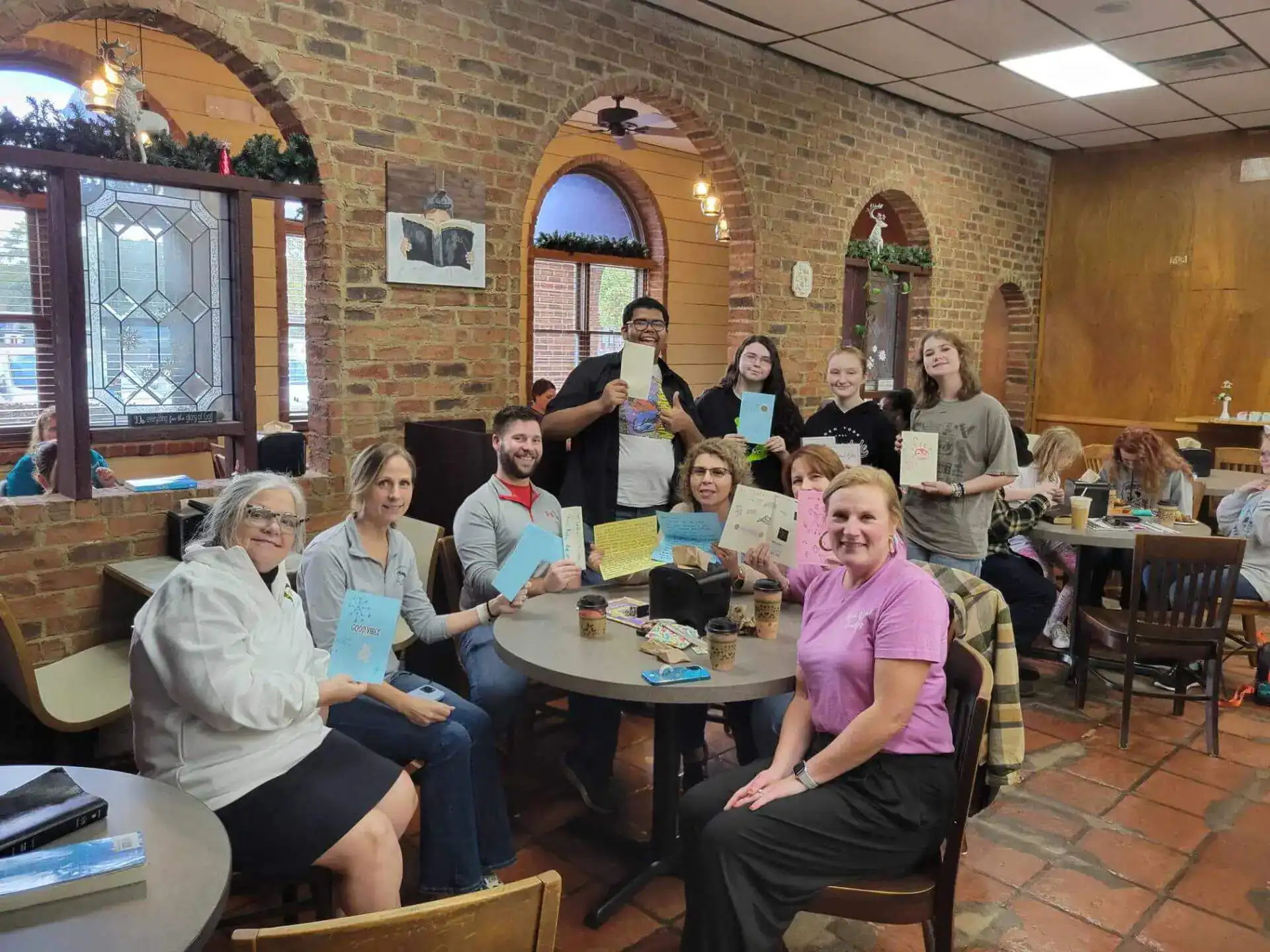 A group of people sitting and standing in a cozy cafe with brick walls, holding up handmade greeting cards.