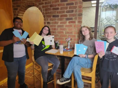 Four people sitting in a cozy café, smiling and holding handmade cards.