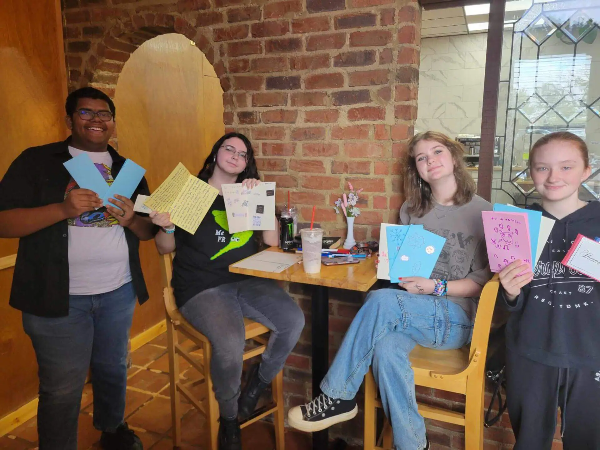 Four people sitting in a cozy café, smiling and holding handmade cards.