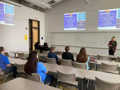 A person stands at the front of a classroom presenting to a small group of seated attendees.