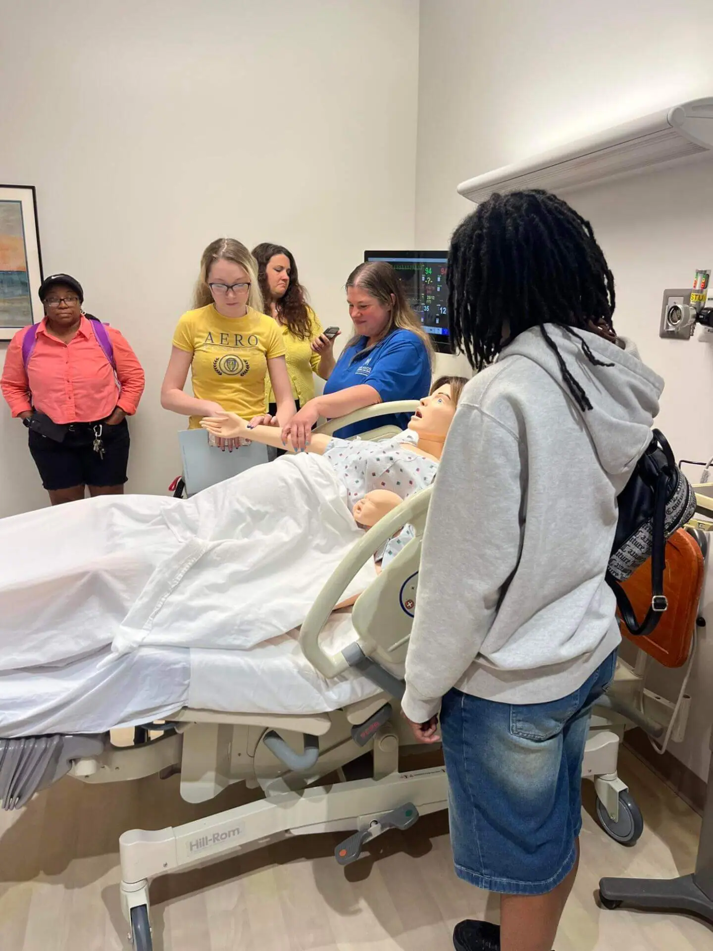 A group of students in a medical training room observe a medical mannequin on a hospital bed.