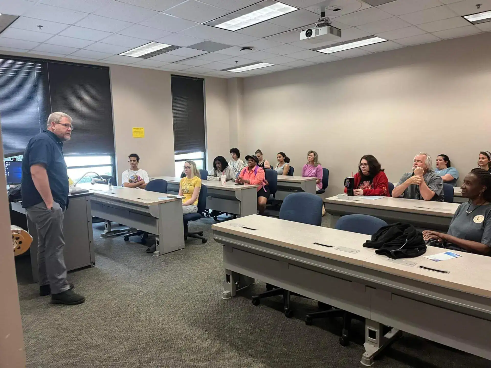A teacher stands at the front of a classroom, engaging a diverse group of attentive students seated at desks with notebooks.