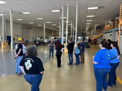 A group of people stands in a spacious industrial facility, listening to a guide.