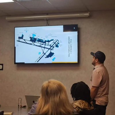 A man in a cap presents an airport capabilities map on a screen to a small group in a meeting room.